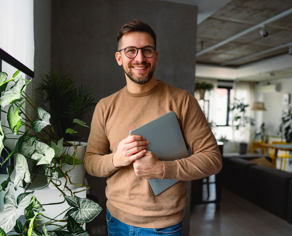 A man holding a laptop smiles in a modern, plant-filled office space.
