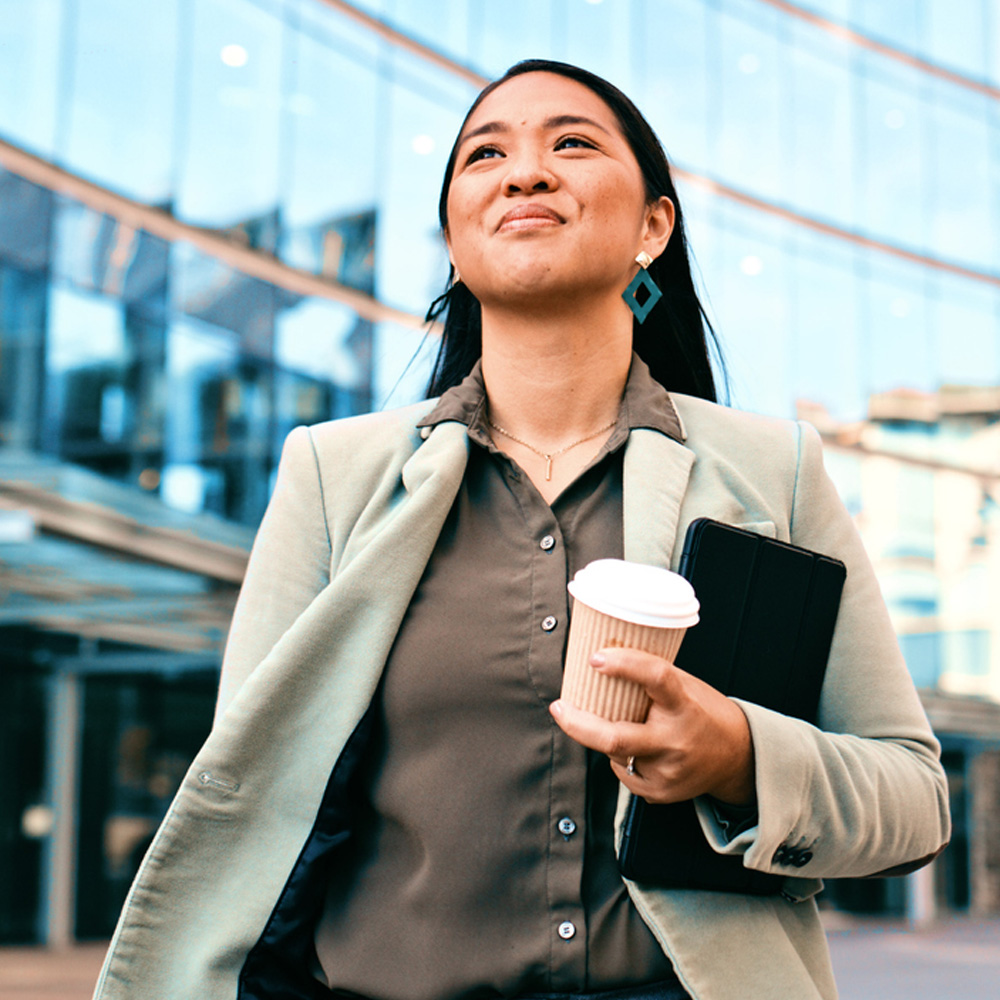 A woman in a blazer holding a coffee cup and tablet outside a modern glass building.
