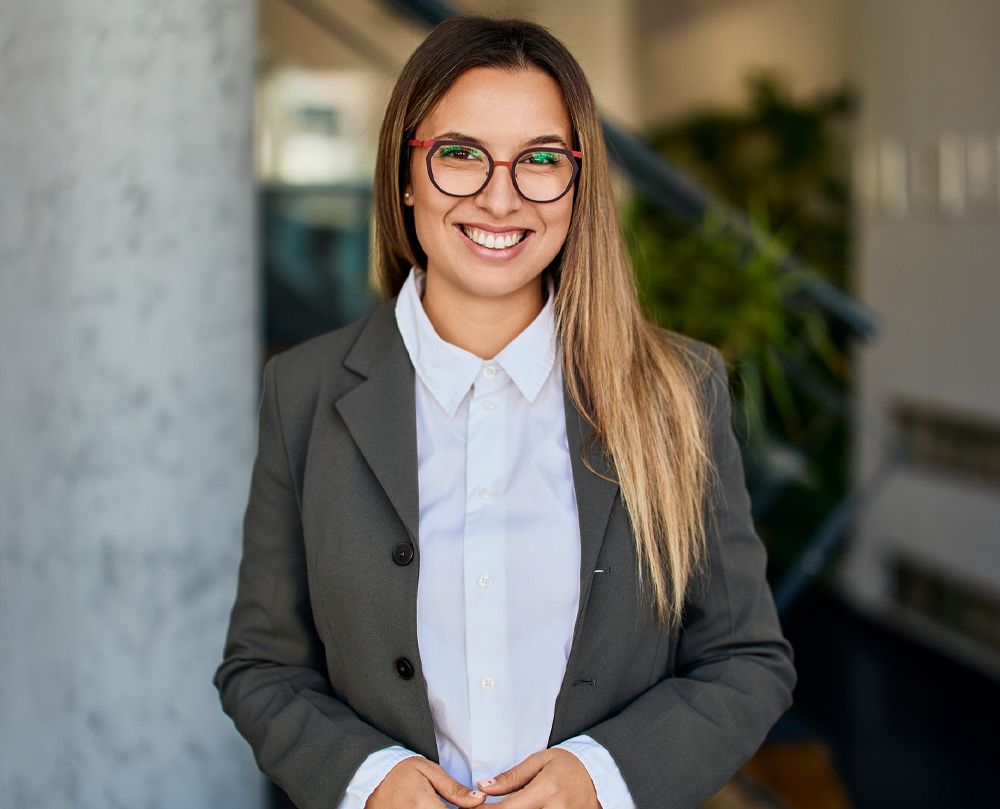 A woman in glasses and a gray blazer smiles confidently indoors.