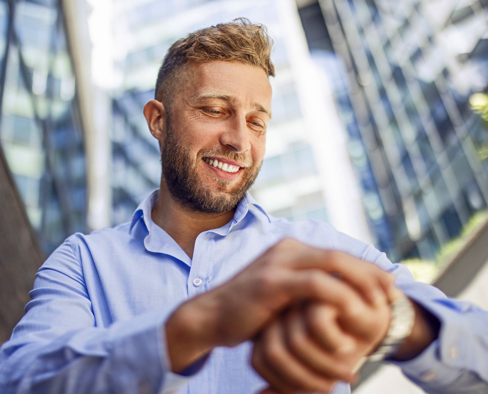 Man in a blue shirt smiling and checking his watch outside skyscrapers.